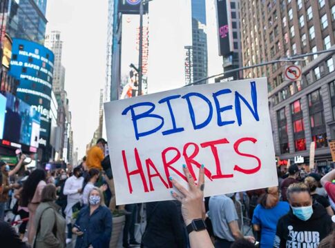 Person holding Biden Harris sign in crowded city street.