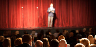 Person on stage speaking to seated audience in theater.
