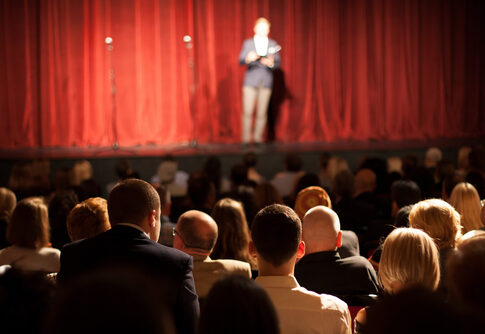 Person on stage speaking to seated audience in theater.
