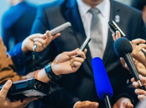 Journalists holding microphones interview a person in a suit.