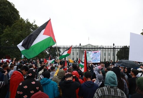 Crowd protesting with Palestinian flags outside a white building.