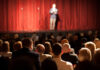 Person on stage speaking to seated audience in theater.