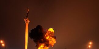 Large blaze in city square near tall monument at night.