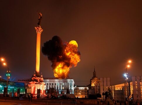 Large blaze in city square near tall monument at night.