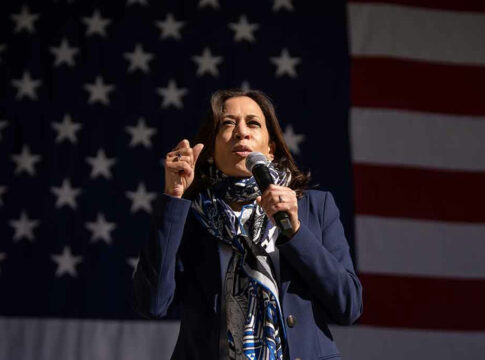 Person speaking into microphone with American flag background.