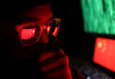 Person wearing glasses with a reflection of the Chinese flag, surrounded by computer screens